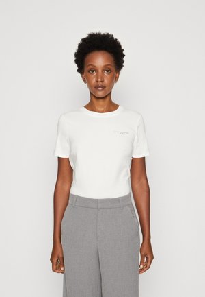 T-shirt bianca a maniche corte con scollo rotondo, caratterizzata da un piccolo logo stampato sul lato sinistro. Indossata con pantaloni grigi su misura.