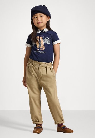 Μαύρη γραφική μπλούζα με άσπρο γιακά, χακί πτυχωτό παντελόνι και καφέ παπούτσια με πολύχρωμες λουρίδες. Παιδί που φορά μπλε μπερέ.