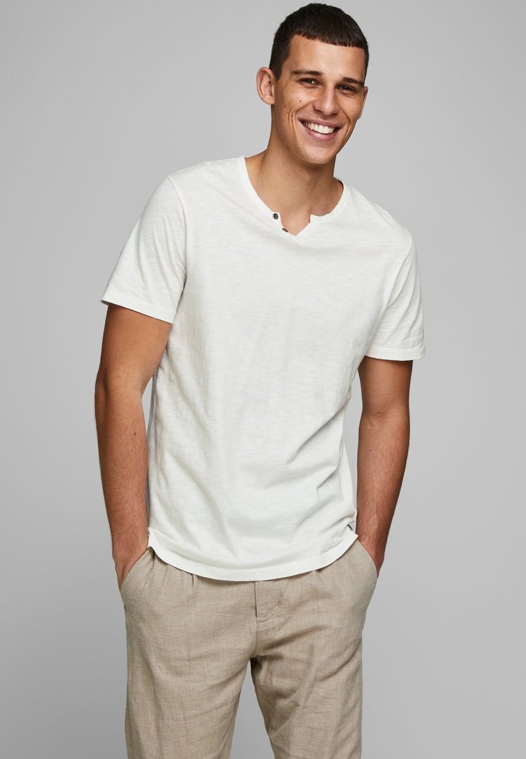 Witte t-shirt met korte mouwen en een ronde halslijn, voorzien van een enkele zwarte knoopdetail, gecombineerd met lichtbeige linnen broek.