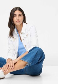 Witte denimjacket met gouden knopen, lichtblauwe top, wijde blauwe jeans en witte sneakers, zittend op een lichte ondergrond.