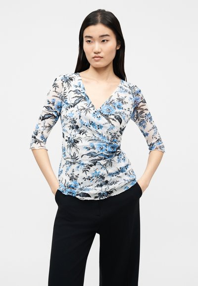 Γυναίκα με μακριά σκουρόχρωμα μαλλιά που φοράει λευκό τοπ τύπου wrap με μπλε ανθισμένο μοτίβο και μανίκια τριών τετάρτων, σε συνδυασμό με μαύρο παντελόνι, με τα χέρια στις τσέπες.