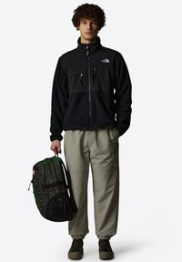Black fleece jacket with a zip front, two chest pockets, and gray pants. A green and black patterned backpack is held in one hand.