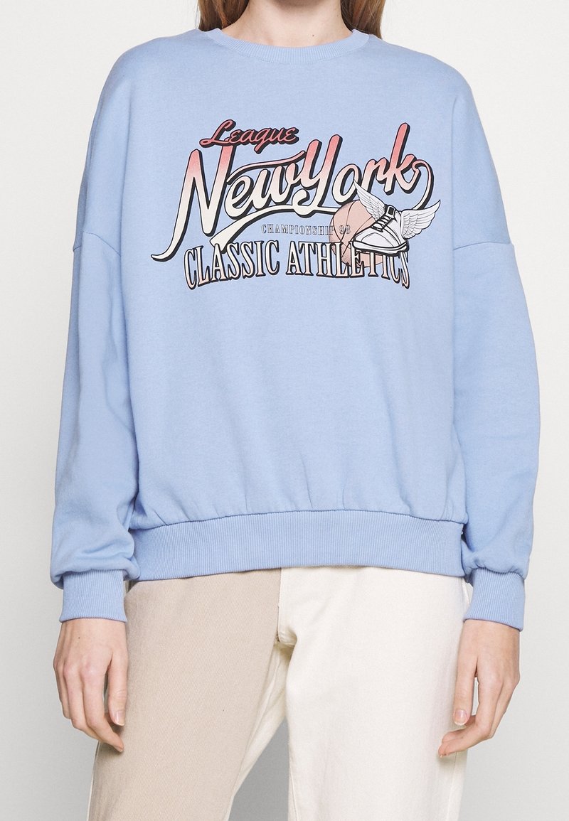 Blå sweatshirt med sänkta axlar, ribbad mudd och midja. Har en djärv grafisk design med "New York" och atletiska teman.