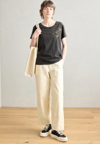 T-shirt grigio scuro con motivo a occhio, pantaloni larghi color crema e sneakers nere con suola bianca. Pavimento in legno naturale e sfondo chiaro.