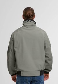 Graue Softshell-Jacke mit hohem Kragen, schwarzen Akzenten und elastischen Bündchen. Verfügt über eine rückseitige Naht und eine glatte, matte Textur.