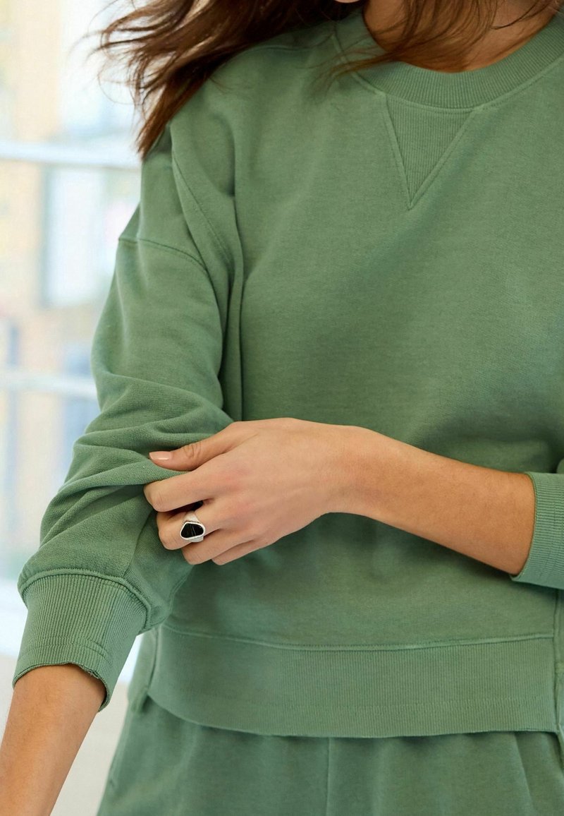 Femme retroussant la manche d’un sweat-shirt vert, portant une bague en argent avec une pierre noire à son doigt.