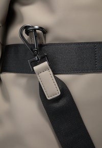 Siv torba, odporna na vodo, z črnim spletom trakom in črnim kovinskim kaveljcem. Ima gladko teksturo in kontrastne poudarke na traku.