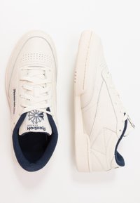 Zapatillas de piel blanca con suela texturizada, detalles en azul marino y perforaciones. Cuentan con cordones y un logo en la lengüeta.