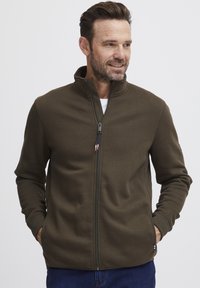 Olivnozeleni zip-up jopič izdelan iz mehkega materiala, z visokim ovratnikom, stranskimi žepi in kontrastnim zadrgo z modrim in rdečim povodcem.