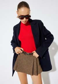 Blazer de mezcla de lana en navy, suéter rojo de canalé, falda de cuadros houndstooth marrón con pliegues y cinturón marrón. Grandes gafas de sol negras con acentos dorados.