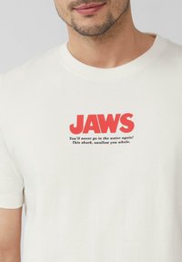 Biała bawełniana koszulka z wyraźnym czerwonym napisem "JAWS" oraz dodatkowym czarnym tekstem poniżej. Standardowy okrągły dekolt i krótkie rękawy.