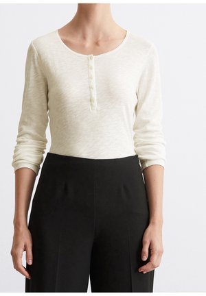 Mujer con una camisa henley de manga larga color crema con botones y pantalones negros de talle alto contra un fondo liso.