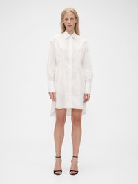 Abito camicia bianco a maniche lunghe realizzato in tessuto leggero, dotato di chiusura con bottoni frontale, colletto appuntito e orlo asimmetrico.
