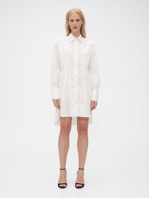 Abito camicia bianco a maniche lunghe realizzato in tessuto leggero, dotato di chiusura con bottoni frontale, colletto appuntito e orlo asimmetrico.