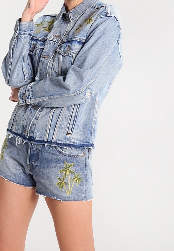 Persona con chaqueta de mezclilla azul claro y pantalones cortos a juego con bordado de palmeras verdes, de pie con los brazos cruzados.