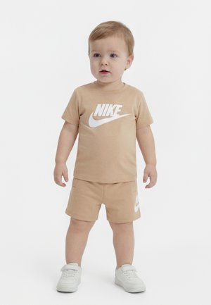 Tout-petit debout portant un t-shirt Nike beige et un short avec des baskets blanches sur un fond clair uni.