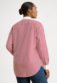 Γυναίκα με σκούρα μαλλιά σε κότσο, που φοράει μακρυμάνικο πουκάμισο με κόκκινες και λευκές κάθετες ρίγες και μπλε τζιν, γυρισμένη πλαγίως σε απλό φόντο.