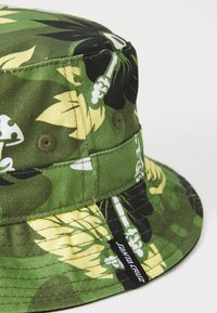 Zeleni platneni šešir s cvjetnim uzorcima u žutoj i crnoj boji, metalnim očicama i crnom etiketom s natpisom "Santa Cruz" sa strane.