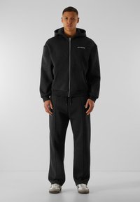 Sweat à capuche noir avec zip, doté de grandes poches avant et de poignets côtelés, associé à un pantalon de jogging assorti avec des coutures latérales. Tissu décontracté avec une texture douce.