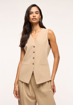 Donna con lunghi capelli scuri che indossa un gilet beige senza maniche con bottoni e pantaloni ampi abbinati su uno sfondo chiaro.