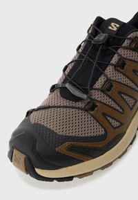 Trail running sko med overdel i brun mesh og sorte detaljer, fleksibel design, beskyttende gummi tåhætte og et sikkert snørresystem.