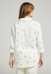 Chemise en coton blanche, texturée, avec broderies florales jaunes et vertes. Elle dispose d'un col et de manches retroussées, ainsi que de coutures décoratives au dos.