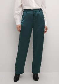 Teal satinbyxor med rak passform, sidfickor och en mjuk textur, tillsammans med en vit skjorta med knappar och svarta ankelstövlar.