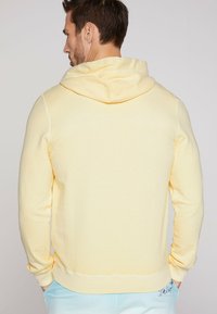 Sudadera con capucha amarilla de acabado texturizado, con puños de canalé y un bolsillo tipo canguro. Presenta costuras de contraste a lo largo de las costuras.
