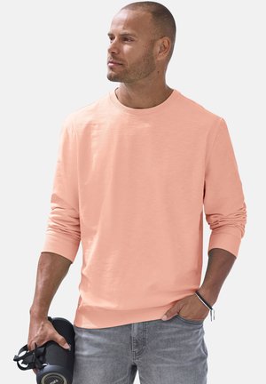 Mann in einem pfirsichfarbenen Langarmhemd und grauer Jeans hält einen schwarzen tragbaren Lautsprecher in der linken Hand, die rechte Hand in der Tasche, schaut nach links.