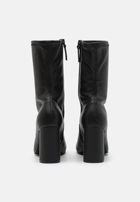 Botas de tobillo de cuero negro con textura suave, tacones altos en bloque y cremalleras traseras para el cierre. Presentan una silueta ajustada y elegante.