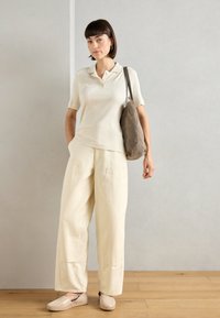 Lichtbeige geribbelde polo, crèmekleurige wijdvallende broek, beige instappers en een grijze schoudertas. Helle texturen en een ontspannen pasvorm.