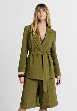 Femme portant un blazer texturé vert olive avec une ceinture et un short large assorti, accessoirisée avec des bracelets au poignet gauche.