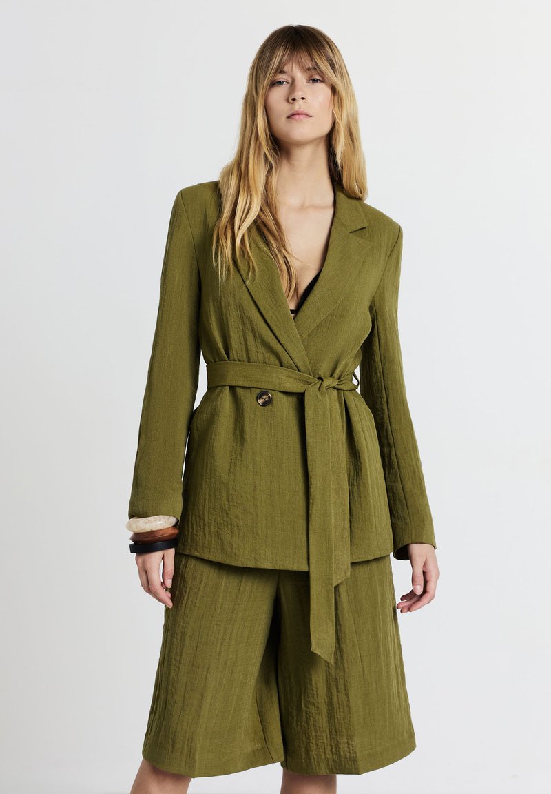 Donna che indossa un blazer testurizzato verde oliva con cintura e pantaloni larghi coordinati, abbinati a braccialetti sul polso sinistro.
