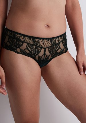 Femme portant des sous-vêtements noirs en dentelle transparente à motif floral, debout devant un fond clair uni.