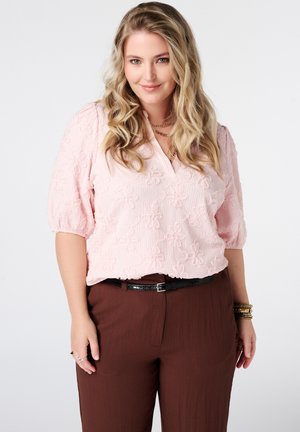 Femme aux longs cheveux blonds ondulés portant un chemisier rose clair texturé avec des motifs floraux et un pantalon marron taille haute avec une ceinture noire.