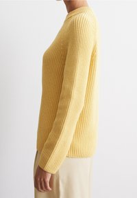 Persona che indossa un maglione lavorato a maglia di un giallo pallido con maniche lunghe, abbinato a pantaloni beige chiaro, mostrata di profilo su uno sfondo bianco.