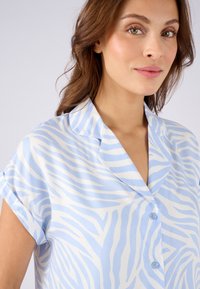 Chemise à manches courtes bleu et blanche avec un motif de rayures de zèbre, dotée d'un col en V et de boutons, fabriquée en tissu lisse.