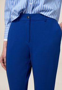 Niebieskie dopasowane spodnie o gładkiej teksturze, z przednią kieszenią i zapięciem na metalowy guzik. W zestawie z jasnoniebieską koszulą w paski.