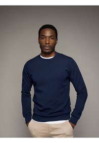 Maglione girocollo blu navy con polsini e fondo a coste. Tessuto morbido, indossato sopra una camicia bianca. Modello in piedi con le mani nelle tasche.