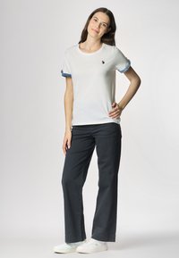 T-shirt bianco in cotone con maniche frangiate blu, caratterizzato da un piccolo logo. Abbinato a pantaloni larghi blu scuro e sneaker bianche.