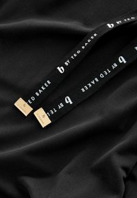 Crna tkanina s zlatnim metalnim detaljima i etiketom koja glasi "b BY TED BAKER" u bijelom tekstu. Dizajn ima glatku teksturu.