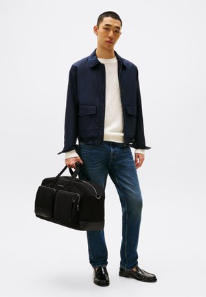 Black duffel bag with two front pockets, smooth texture, and contrasting lower accents. Model wears a navy jacket and cream knit sweater.