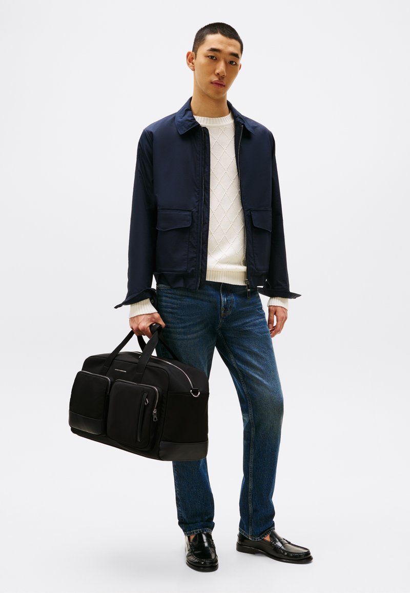 Sac de sport noir avec deux poches avant, texture lisse et accents inférieurs contrastés. Le modèle porte une veste marine et un pull en tricot crème.