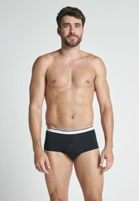 Svarta bomullsboxer med vit elastisk midjeband som har varumärkesnamnet. Modellen har ljus hudton och kort mörkt hår.