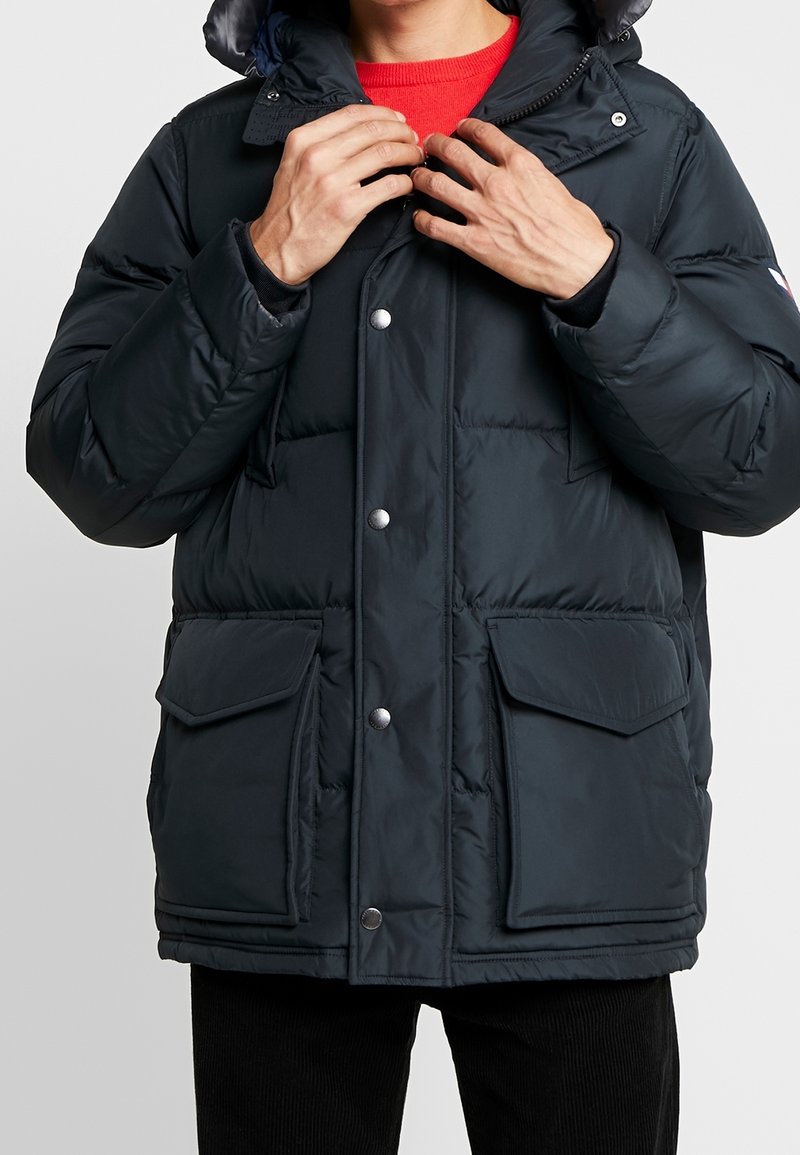 Tmavě námořnická prošívaná bunda s kapucí, opatřená patentními knoflíky, dvěma předními kapsami a prošívanou texturou pro izolaci.