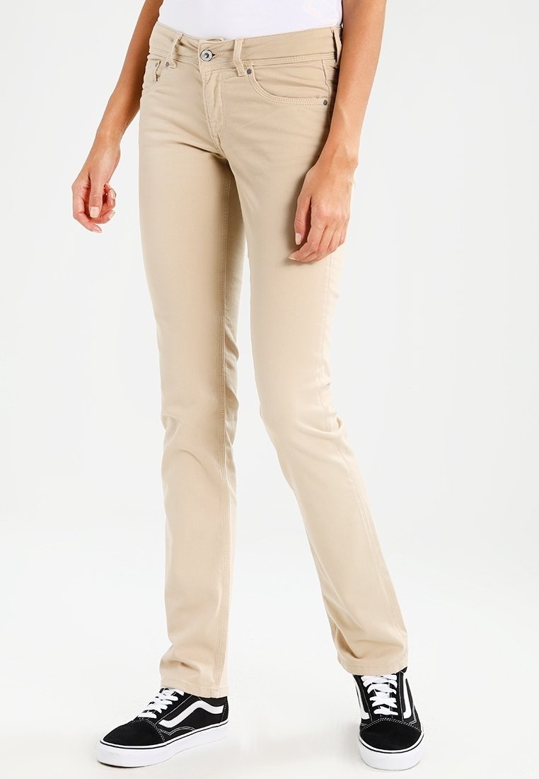 Femme portant un pantalon beige slim et des baskets noires et blanches, debout sur un fond blanc uni.