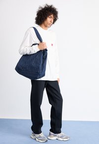 Navy tote bag with white stripes and dots, made of fabric, featuring a slouchy design. Worn over the shoulder by a person in a white long-sleeve shirt.