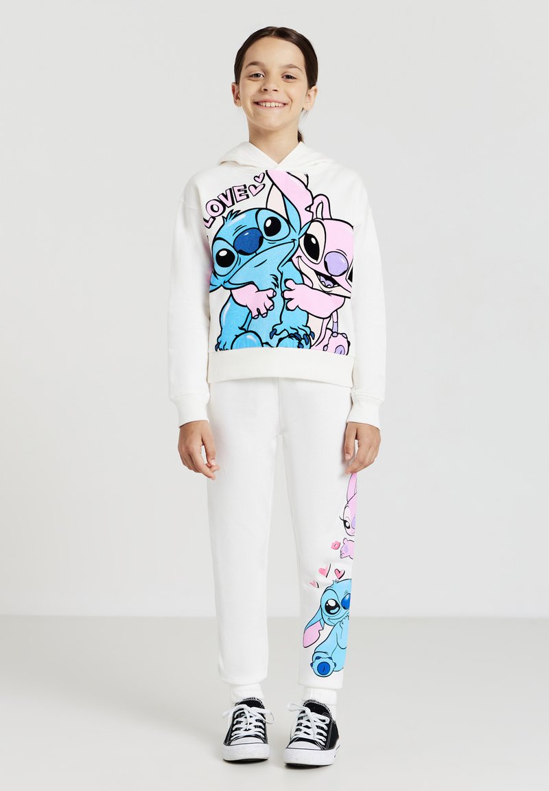 Felpa bianca con personaggi dei cartoni animati blu e rosa, testo "LOVE". Pantaloni della tuta bianchi con disegni rosa e blu. Scarpe da ginnastica nere.