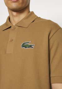 Ruskea polo-paita, jossa on teksturoitu kangas, sisältäen vihreän ja valkoisen brodeeratun krokotiililogon sekä kaksi nappia kauluksessa.