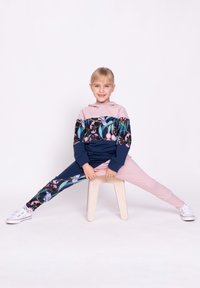 Dziecko w bluzie z kapturem z blokami kolorów z kwiatowym wzorem w granacie i różu, w połączeniu z różowymi spodniami. Siedzi na jasnym, drewnianym stołku.